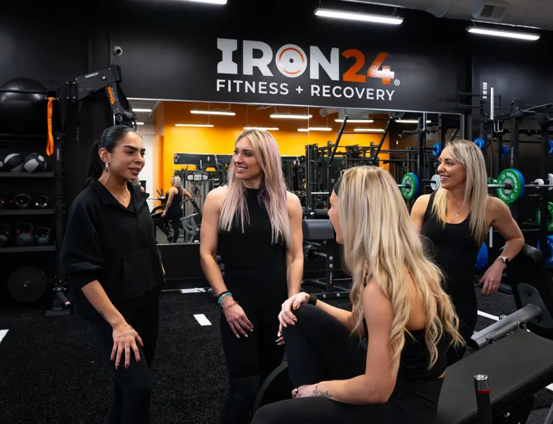 Four Iron 24 coaches laughing together on the gym floor beneath the Iron 24 Fitness + Recovery logo