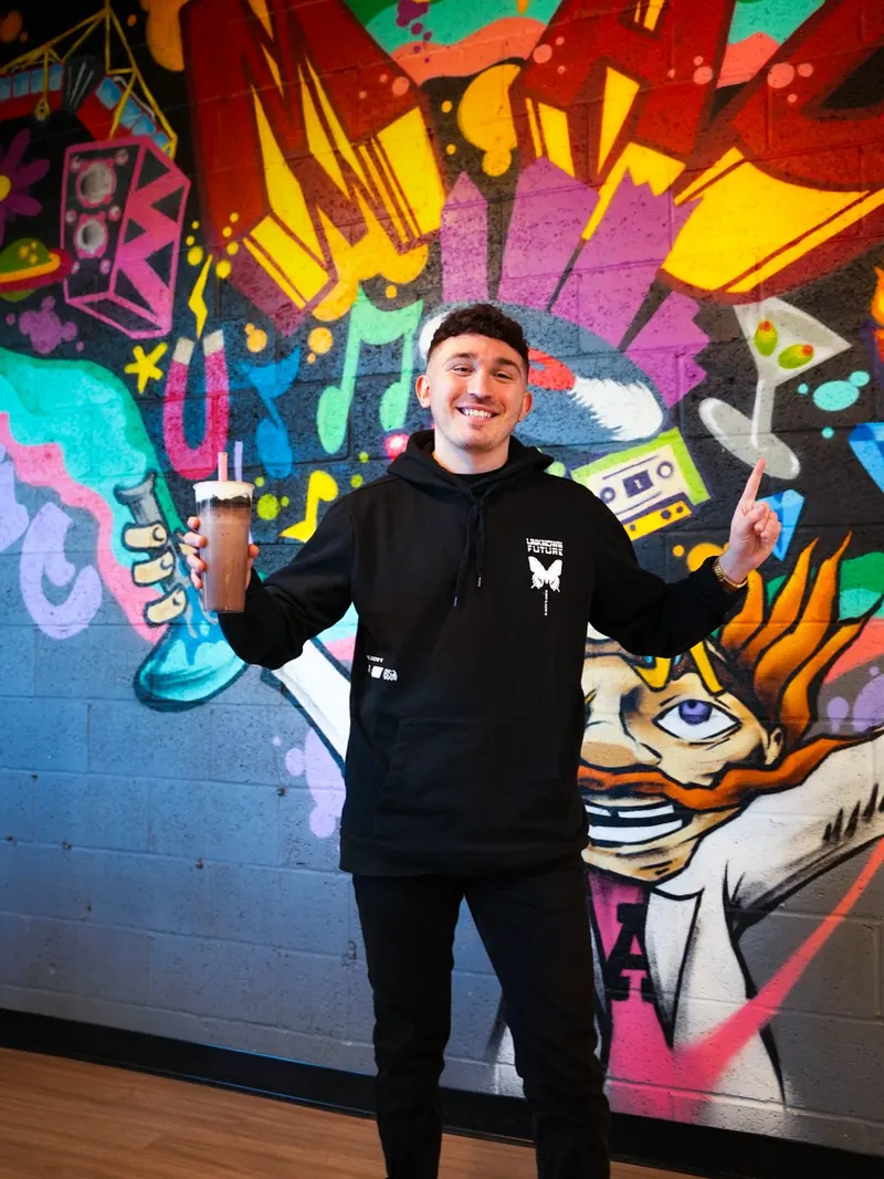 Mike grinning in front of the graffiti mural, bubble tea in one hand and the other pointing skyward