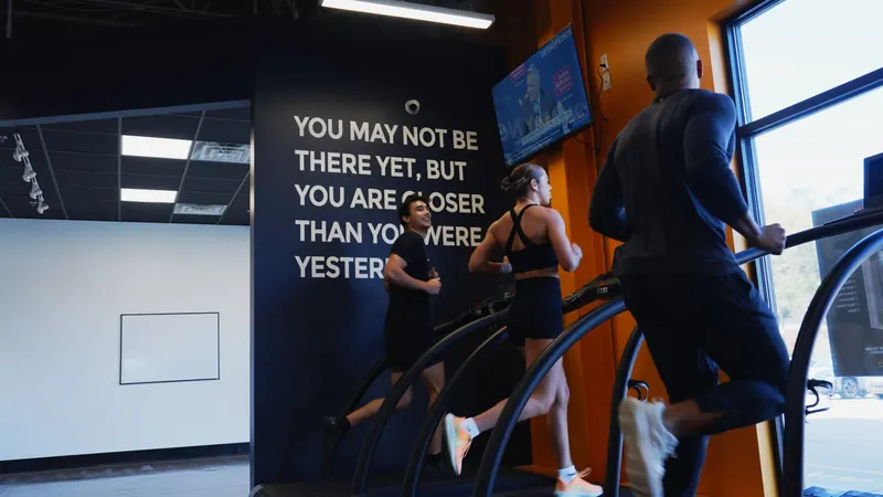 Two members mid-stride on stair climbers beneath the quote wall reading 'you may not be there yet, but you are closer than you were yesterday'