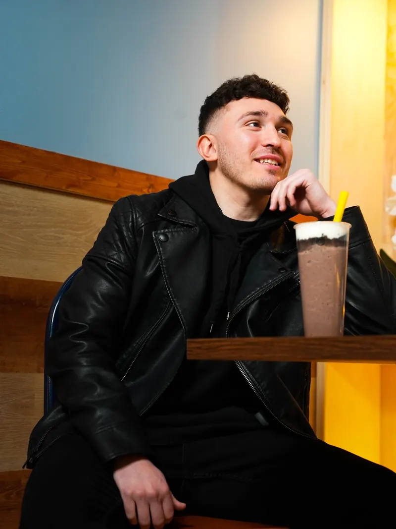 Mike in a leather jacket at a table, chin on his hand, smiling off-camera beside a chocolate bubble tea with a yellow straw