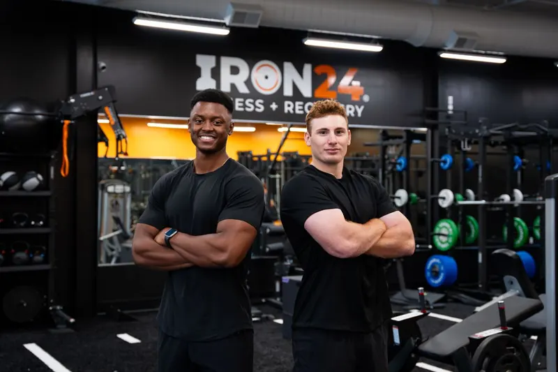 Two Iron 24 trainers in front of the gym's logo wall