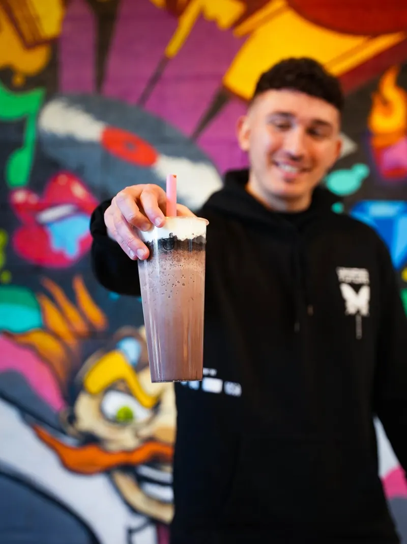 Creator in a black hoodie holding a chocolate bubble tea out toward the camera in front of Dr. Tea's graffiti mural
