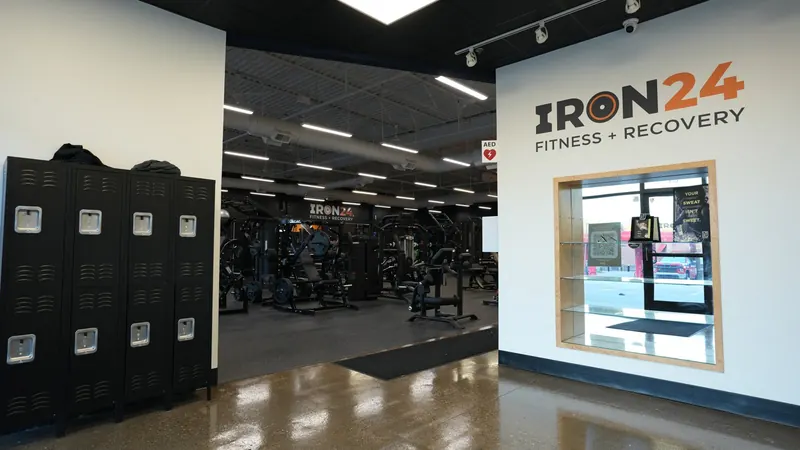 Black lockers and the Iron 24 Fitness + Recovery logo wall at the gym entrance, with an empty equipment floor behind them