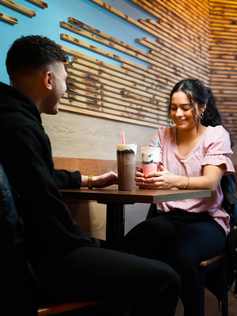Emily Harrigan and Mike DeSabato laughing across a table over two bubble teas beneath Dr. Tea's slatted wood wall